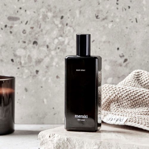 En svart flaska märkt 'Meraki room spray' på en grå yta, bredvid en handduk och en liten behållare, mot en betongvägg.