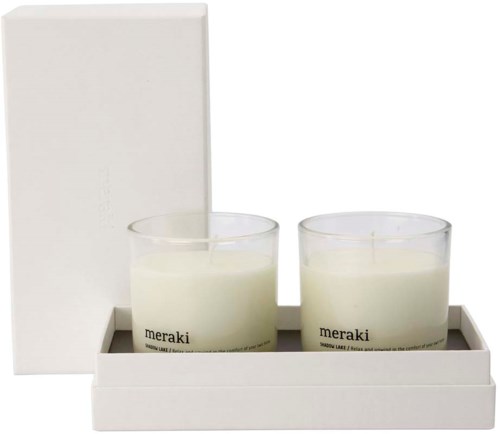 Två vita ljus i glasvaser med 'meraki'-etiketter, placerade i en vit bricka med en låda bakom.