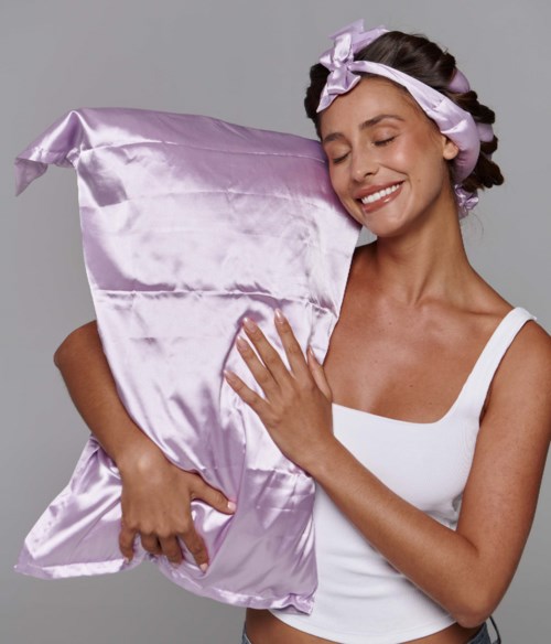 En smilende kvinne i en hvit tanktop holder en stor lilla satinpute, iført en satinbånd i håret.