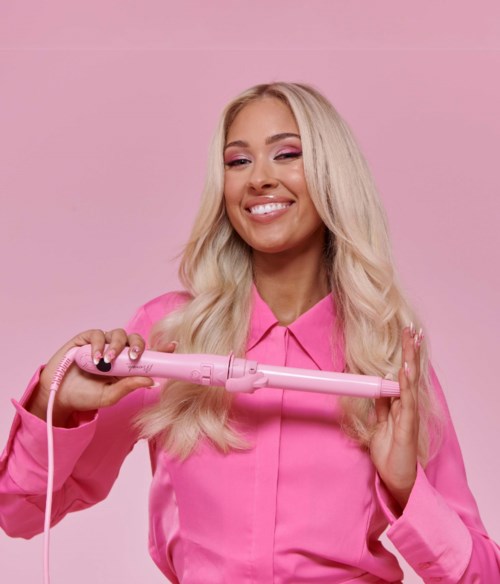 En kvinna med långt blont hår i rosa kläder lockar håret med ett rosa lockverktyg.
