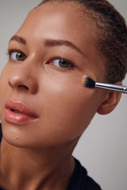 En kvinna med klart hud har makeup appliceras med en liten borste på kinden. Hon ser lugn och avkopplad ut.
