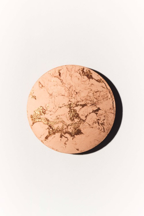 En rund, rosa-beige makeup-puder med guldmarmering på ytan, placerad på en vit bakgrund.