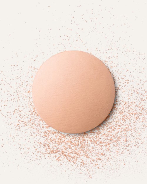 En persikfärgad makeup-svamp med rosa puder utspritt runt den på en vit bakgrund.