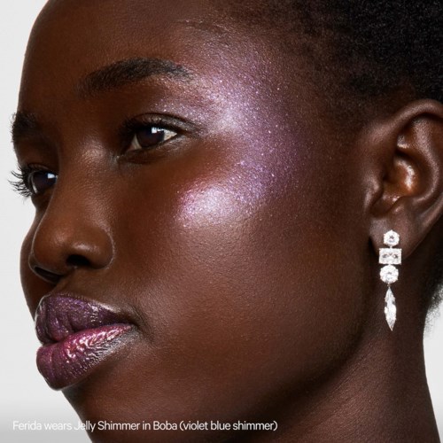A woman with dark skin wears shiny purple lip gloss, sparkly purple eyeshadow, and earrings. Her hair is short and styled.