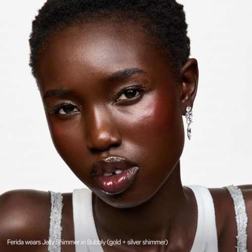A woman with dark skin and natural hair, wearing silver earrings and a tank top. She has shiny lips and glowing skin.