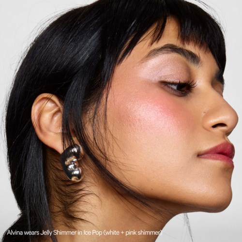 A woman with dark hair and earrings wears makeup and a shiny lip color, looking sideways with a confident expression.