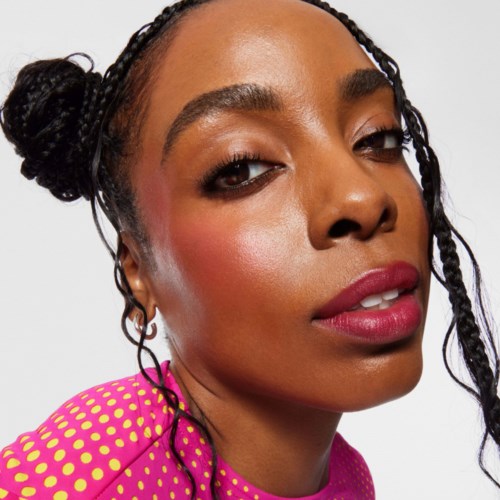 En kvinde med fletninger, makeup, i en pink prikketop, ser selvsikkert på kameraet.