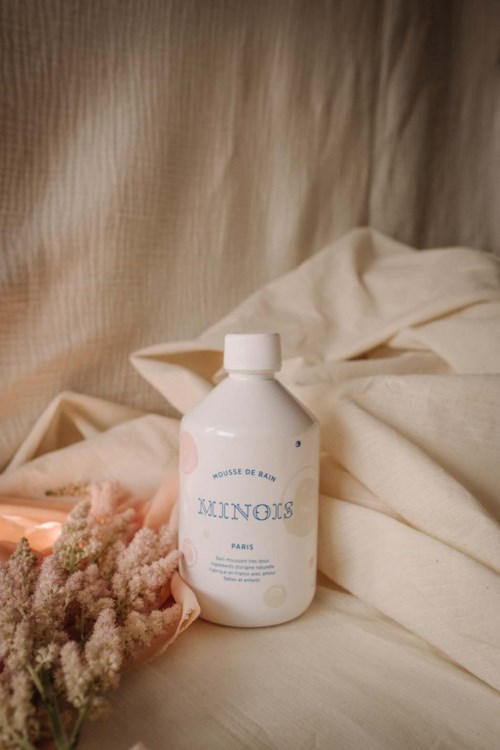 Een witte fles met "Miniols" op een stoffen oppervlak, met zachte stof en roze bloemen in de buurt.