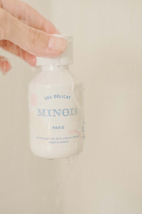 En hand håller en liten vit flaska märkt 'MINOIS PARIS' med blå text. Flaskan verkar innehålla en gel.