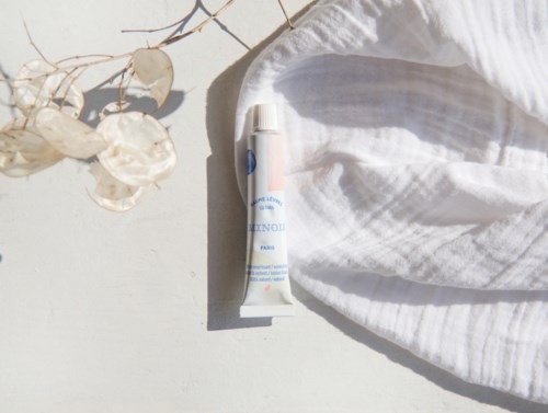 A tube of ointment, white cloth, and dried plants on a light surface with sunlight and shadows.