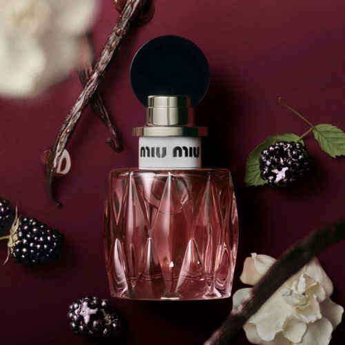 Miu Miu parfymespray med sort top på mørkerød baggrund omgivet af bær og hvide blomster.