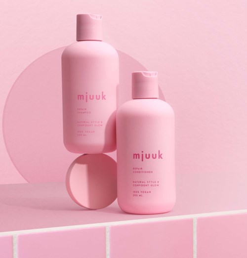 To rosa flasker merket 'mJUk' står på en rosa fliseoverflate, med en rosa sirkel i bakgrunnen.