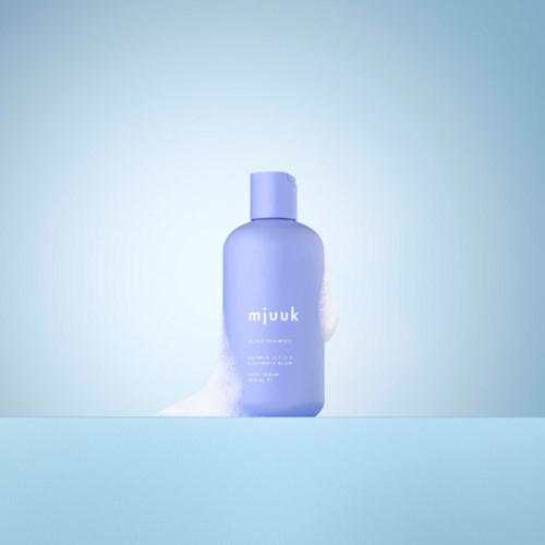 En lilla flaske med hudpleieprodukt kalt 'mjuuk' står på en overflate med skum ved siden av, mot en myk blå bakgrunn.