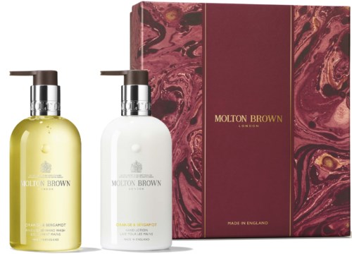 Kaksi Molton Brown käsisaippuapulloa, yksi keltainen ja yksi valkoinen, taustalla vaaleanpunainen ja kultainen rasia.