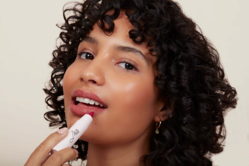 A woman with curly hair applies lipstick and looks at the camera with a soft smile.