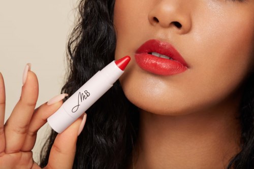 A woman applies red lipstick using a lipstick-shaped device with 'MsB' written on it.