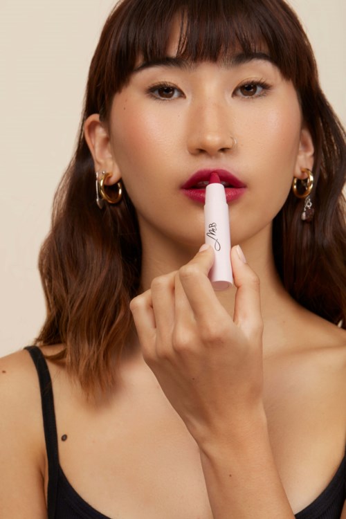 A woman with brown hair and earrings is applying lipstick. She wears a black top. The background is plain.