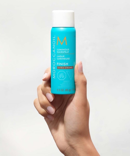 Eine Hand hält eine blaue Flasche Moroccanoil Luminous Hairspray vor einem weißen Hintergrund.