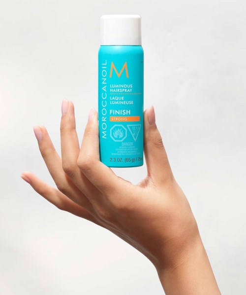 En hand håller i en blå flaska Mochaccino Luminous Hairspray med vit kapsyl, mot en enkel bakgrund.