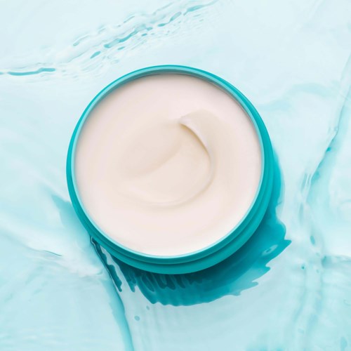 En rund beholder med hvid creme eller lotion på en blå overflade, med noget spildt creme omkring.