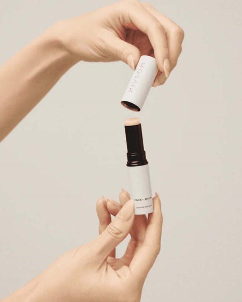A person is opening a white bottle, revealing a stick-like object inside. The background is plain and light-colored.