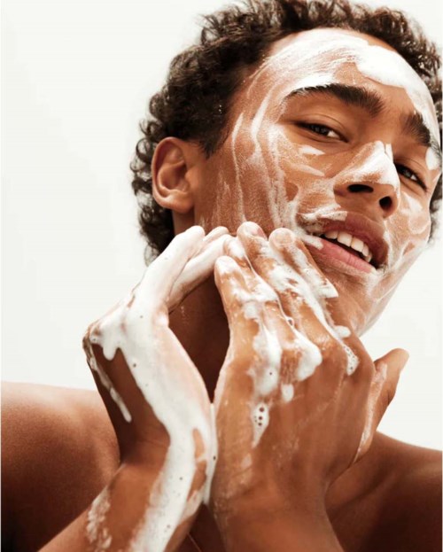 A young man with curly hair cleans his face with soap, smiling and looking confident.
