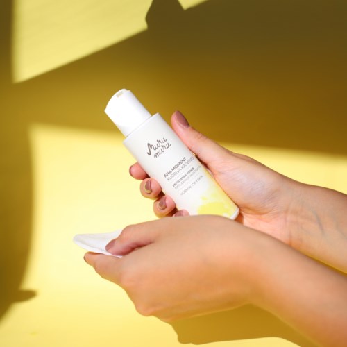 Hands holding a white bottle with a yellow label against a yellow background. The bottle has a white cap.