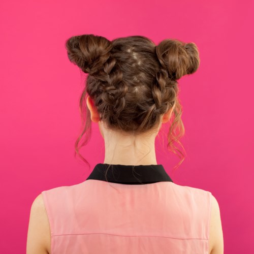 A girl with two braided buns on pink background, wearing a sleeveless pink top and a black collar.