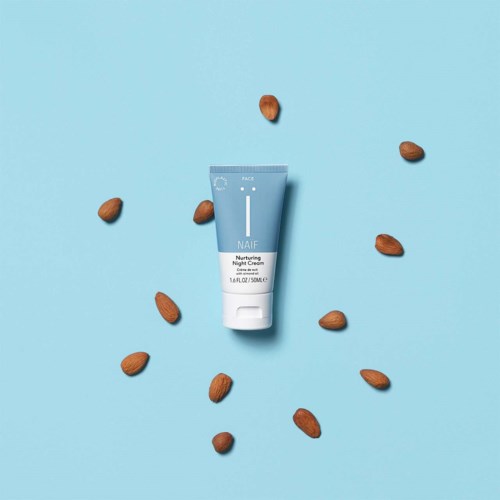 En blå tub märkt 'Nurturing Night Cream' omgiven av utspridda mandlar på en blå bakgrund.