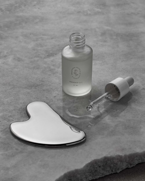 A skincare serum bottle, a dropper with serum, and a ceramic heart-shaped dish. They are on a grey surface.