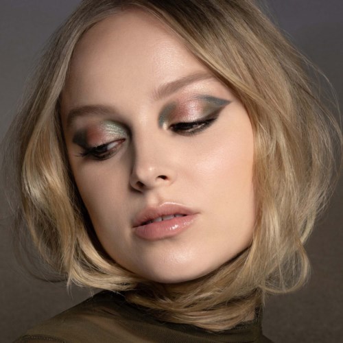 En kvinne med kort blondt hår og elegant øyemakeup ser nedover, med en nøytral bakgrunn.