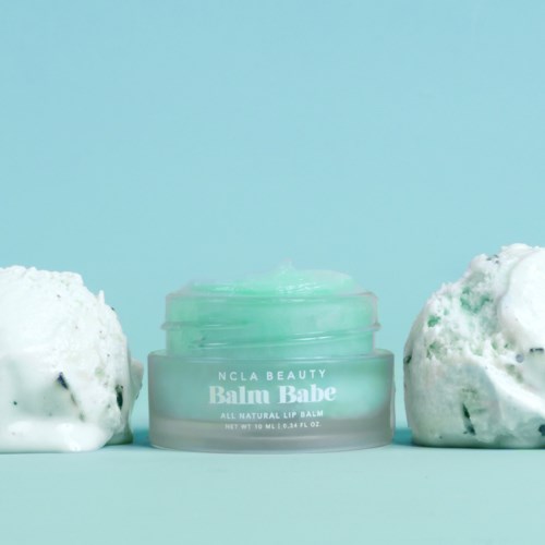 En liten grön burk märkt 'Balmy Babe All Natural Lip Balm' med två vita fluffiga föremål på en blå bakgrund.