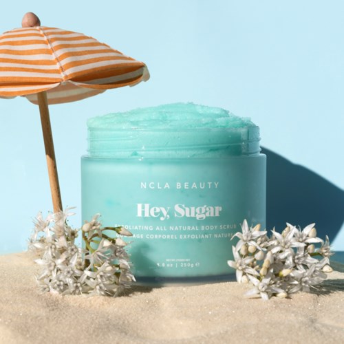 A light blue container of body scrub with a beach umbrella and white flowers under bright sunlight.