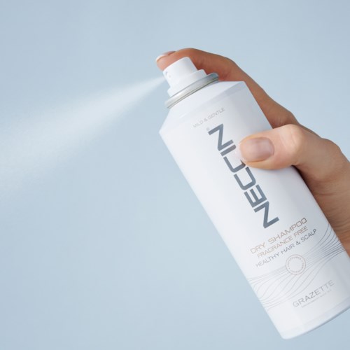 En hånd sprayer en hvid dåse af NeoCos tørshampoo til hår og hovedbund. Baggrunden er lyseblå.