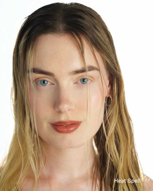 Een jonge vrouw met blond haar, blauwe ogen en rode lippen, glimlacht zachtjes met een neutrale achtergrond.