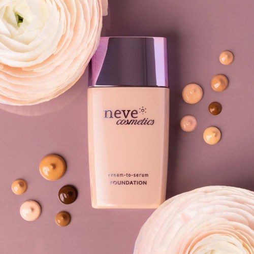 En flaska foundation från Neve Cosmetics omgiven av små, runda pigmentprover och två stora vita blommor.