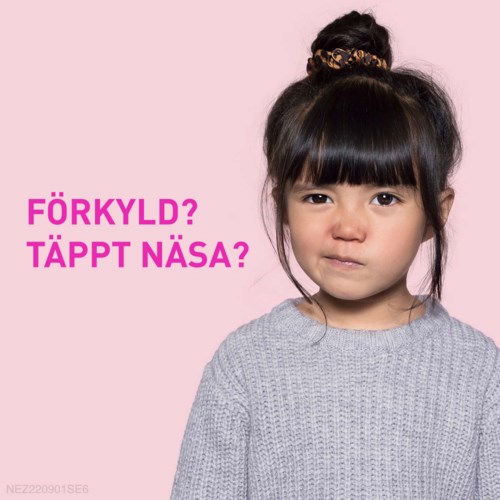 En ung flicka med mörkt hår i en knut och lugg, bär en grå tröja, tittar mot kameran med ett svagt leende.