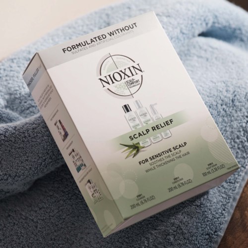 En låda med Nioxin produkter för känslig hårbotten, med flaskor och ett grönt aloe blad på vit och grön förpackning.