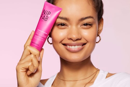 En smilende kvinde holder et pink tube med hudplejeprodukt kaldet NIP+FAB mod en pink baggrund.