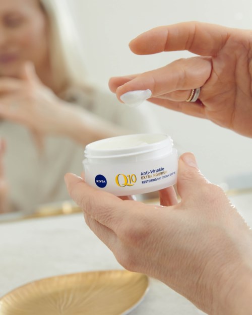 Någon applicerar ansiktskräm, med en kvinna i bakgrunden. Ansiktskrämen är Nivea Q10 Anti-Wrinkle.