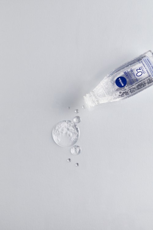 En liten flaska märkt 'AQUA' spiller vatten på en vit yta och bildar droppar och en pöl.