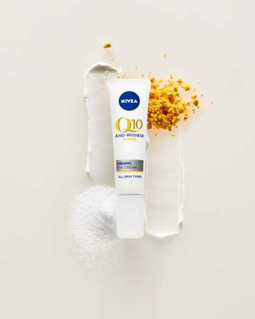 Nivea Q10 Anti-Wrinkle Fuktighetskräm för ögon med vita och gula element, omgiven av pulver på ljus bakgrund.