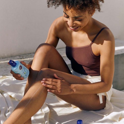 A young woman with curly hair applies sunscreen on her leg, smiling, while sitting on a white cloth.