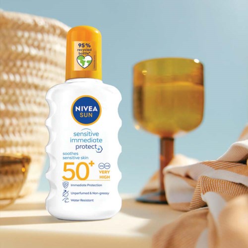 A white bottle of Nivea Sun sunscreen with 50+ protection, on a table with a lamp and cloth, sunny background.