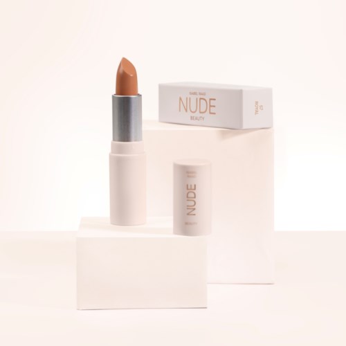 Een nude lippenstift in een wit doosje, met een doos achter en een kleine dop in de buurt.