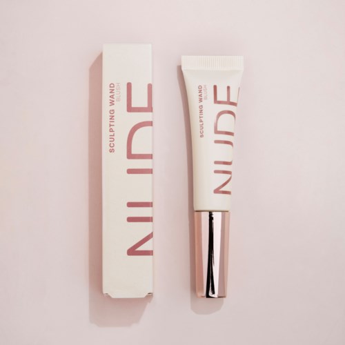 En hvit tube og eske merket 'NUDE' og 'sculpting wand' ligger på en rosa bakgrunn.
