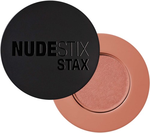 Musta pyöreä rasia, jossa lukee "NUDESTIX STAX," ja se peittää osittain vaaleanpunaisen poskipunan tai luomivärin.