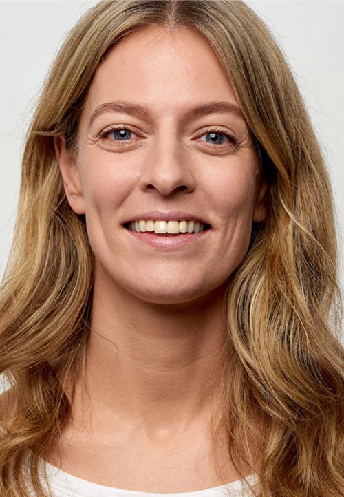 En smilende kvinde med langt blond hår, blå øjne, lys hud iført hvid skjorte.