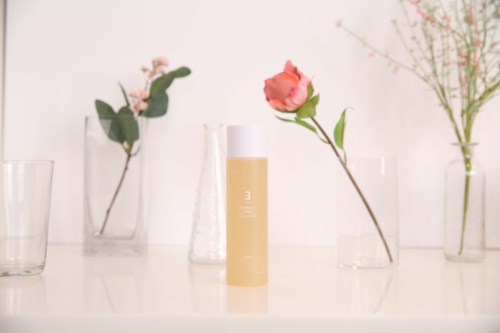A beige bottle is centered on a white surface, surrounded by various clear glass vases with pink and white flowers.