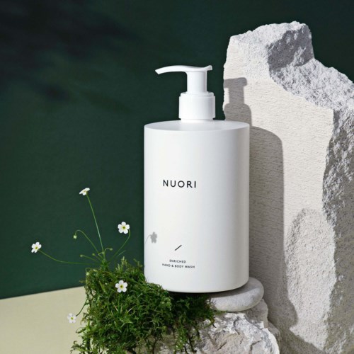 Valkoinen Nuori-vartashampoo pullo, jossa on annostelija, asetettu pyöreälle kivelle, lähellä kukkia ja vihreää, tummanvihreän taustan edessä.
